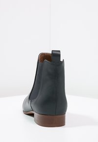 Stivale alla caviglia in pelle nera con suola marrone, pannello laterale elastico e linguetta, mostrato da dietro su superficie bianca.