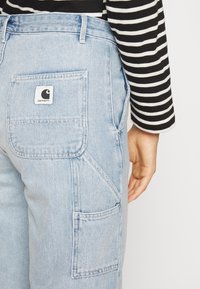 Pantalones cargo de denim azul claro con un bolsillo trasero de parche y una etiqueta de Carhartt. Combinados con una camiseta de manga larga a rayas en blanco y negro.