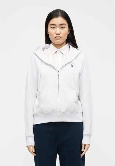 Νεαρή γυναίκα που φοράει λευκό φούτερ με φερμουάρ πάνω από ριγέ πουκάμισο με γιακά και σκούρο μπλε παντελόνι, όρθια μπροστά σε ένα απλό φωτεινό φόντο.