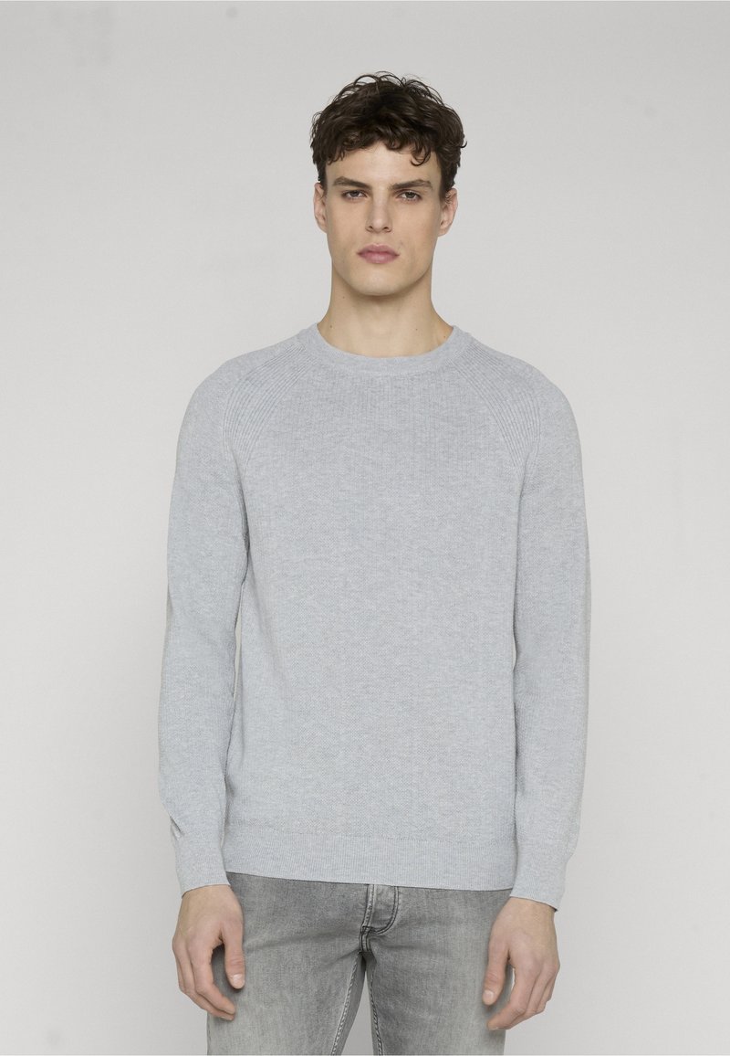Jeune homme aux cheveux courts et bouclés, portant un pull en maille gris clair et un jean gris, debout devant un fond clair uni.