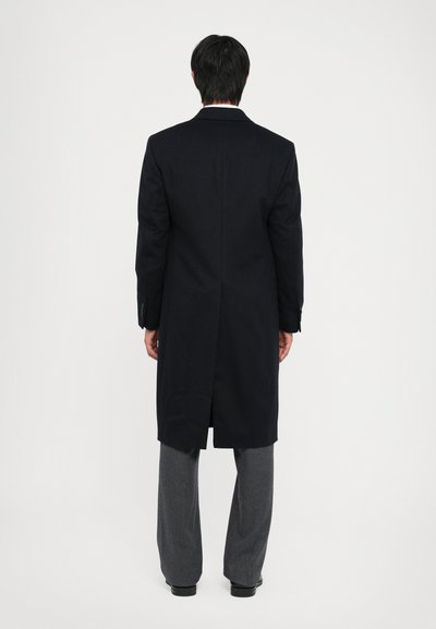 Cappotto di lana blu navy con uno spacco posteriore singolo, maniche lunghe e polsini con bottoni, abbinato a pantaloni grigi su misura.