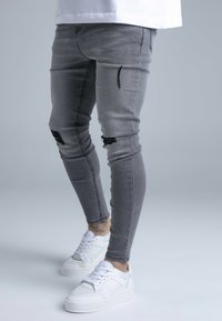 Szürke szűk szabású farmer, szakadt térdekkel, fehér sportcipőkkel párosítva, vastag talppal és texturált felsőrésszel.