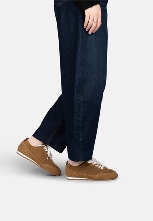 Persona che indossa jeans blu scuro e sneaker in camoscio marrone con lacci bianchi, in piedi con un piede leggermente sollevato.