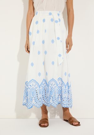 White midi skirt with blue floral and geometric embroidery near the hem, worn with a white top and brown slip-on sandals.