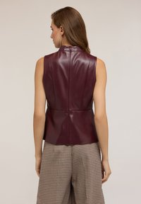 Una donna con capelli lunghi indossa un top senza maniche in pelle bordeaux scuro e pantaloni ampi beige a scacchi, in piedi di spalle contro uno sfondo neutro.