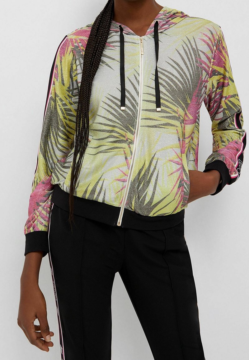 Femme portant un sweat à capuche zippé imprimé de feuilles tropicales jaunes et roses, cordons noirs, pantalon noir assorti avec des bandes latérales, main dans la poche.