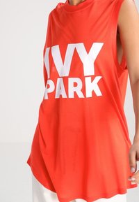 Rød ærmeløs top lavet af letvægtsstof med et fremtrædende hvidt "IVY PARK" logo centreret på forsiden. Normal pasform med rund halsudskæring.