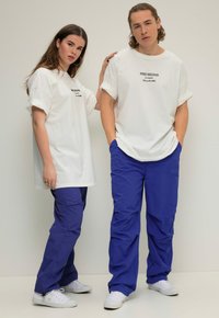 Witte oversized t-shirts met opgerolde mouwen, voorzien van zwarte tekst, gecombineerd met blauwe broeken. Modellen dragen witte sneakers en tonen een relaxte pasvorm.
