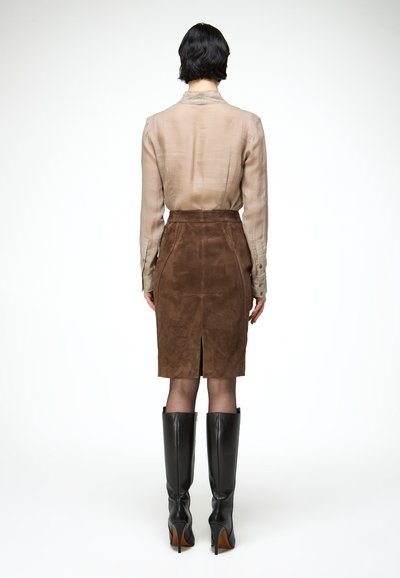 Femme debout de dos, portant un chemisier beige, une jupe en daim marron mi-longue avec une fente au dos, et des bottes longues à talons noirs sur fond blanc.