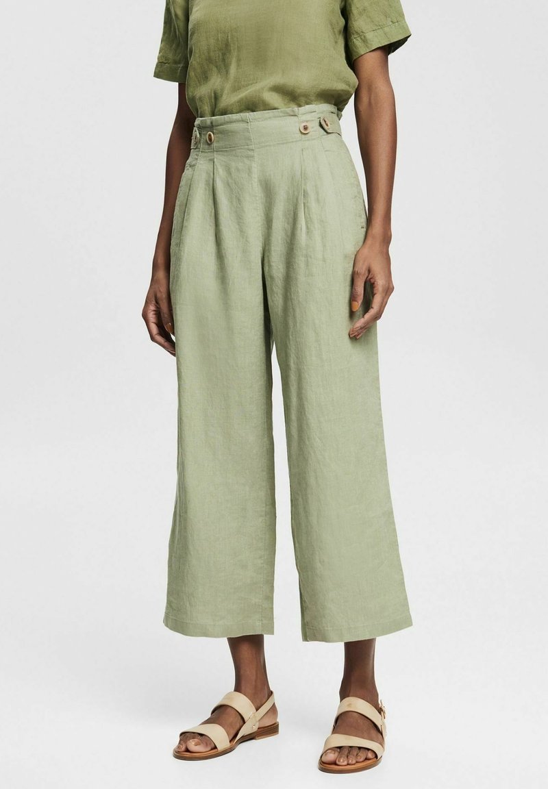 Mujer vistiendo pantalones de lino cortos de pierna ancha en verde claro con detalles de botones en la cintura y sandalias abiertas beige.