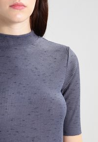 Lyhythihainen harmaa yläosa, tehty ribbineuloksesta, jossa korkea kaula-aukko ja teksturoitu kuvio, jossa satunnaisesti tummempia pilkkuja.