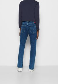 Blå denimjeans med rak ben-design, två bakfickor och en svart midjebandsetikett. Tillsammans med en mörk långärmad topp och vita skor.