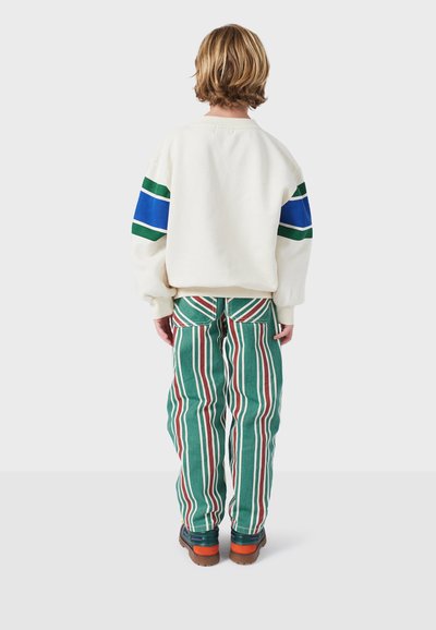 Felpa bianca con strisce verdi e blu; pantaloni a strisce verdi e rosse; scarpe arancioni e blu navy; vista in primo piano da dietro.