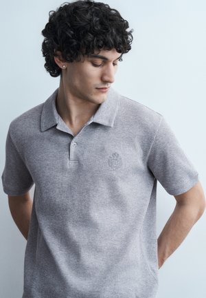 Νεαρός με σγουρά σκουρόχρωμα μαλλιά, φορώντας γκρι ανοιχτό φανελένιο πόλο μπλουζάκι με κοντά μανίκια, κοιτάζοντας προς τα κάτω με τα χέρια πίσω από την πλάτη.