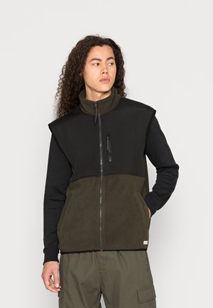 Chaleco de forro polar con parte superior negra y parte inferior verde oliva, que presenta un alto cuello, un bolsillo con cremallera en el pecho y dos bolsillos laterales.