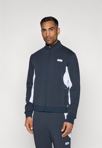 Tmavomodrá zipovaná bunda s bielymi bočnými panelmi, rebrovanými manžetami a nášivkou loga na hrudi. Navrhnutá s športovým strihom.