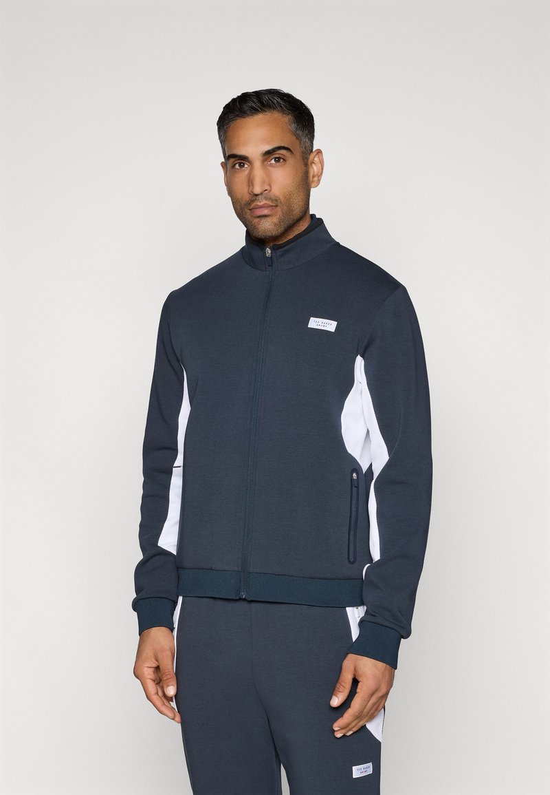 Tmavomodrá zipovaná bunda s bielymi bočnými panelmi, rebrovanými manžetami a nášivkou loga na hrudi. Navrhnutá s športovým strihom.