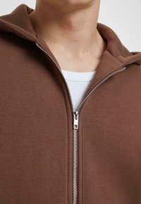 Brauner Reißverschluss-Hoodie aus strukturiertem Stoff, mit einem silbernen Reißverschluss und einer weißen inneren Schicht, die am Halsbereich sichtbar ist.