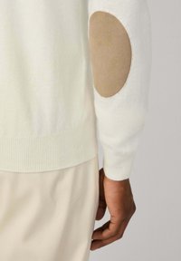 Krupan kadra osobe koja nosi kremasti pulover s bež ovalnim zakrpama na laktovima i krem hlačama, ruka opuštena uz tijelo.