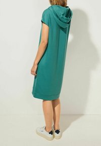 Turkoois katoenen hoodiejurk met korte mouwen, knielengte, met een afgeronde zoom en minimalistisch ontwerp. Gemaakt stijl met witte sneakers.