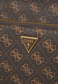 Braune synthetische Handtasche mit einem geprägten Logo-Muster, goldenen Reißverschlussdetails und einer dreieckigen goldenen Logo-Platte mit "GUESS USA".