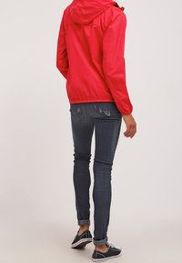 Rode lichtgewicht jas met capuchon, gecombineerd met donkerblauwe jeans met versleten details, en zwarte sneakers met witte zolen.
