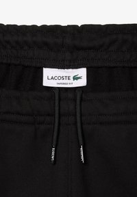 Pantalon de survêtement noir à coupe fuselée avec une taille élastique et des cordons de serrage, arborant une étiquette Lacoste avec un logo crocodile vert.