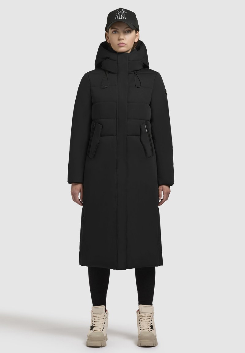 Crna puffer jakna s visokim ovratnikom, prednjim patentnim zatvaračem i dva bočna džepa. Nosena s crnom kapom i svijetlosmeškim čizmama sa vezicama.