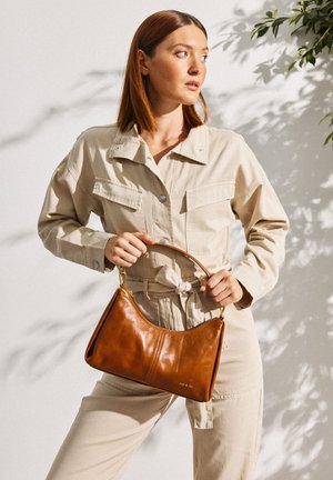 Femme en combinaison beige tenant un sac à main en cuir marron d'une main, debout contre un mur clair avec des ombres douces de plantes.