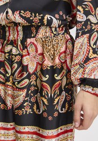 Zwarte stoffen jurk met ingewikkelde rode, gele en witte bloemen- en paisleypatronen, met een elastische taille en gegatherde mouwen.