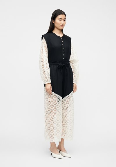 Femme portant une robe noire sans manches avec des manches longues et un ourlet en dentelle blanche, taille cintr ée, debout en talons blancs pointus sur un fond uni.