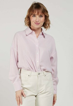 Femme aux cheveux courts et ondulés portant une chemise à rayures roses et blanches, boutonnée et rentrée dans un pantalon taille haute blanc cassé, debout devant un fond uni.