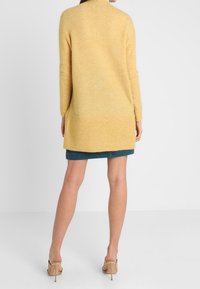 Cardigan lungo giallo a maglia con scollatura rotonda e orlo a coste, abbinato a una gonna in denim blu e sandali beige con punta aperta.