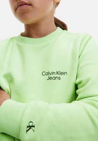 Světlé zelené tričko s logem "Calvin Klein Jeans" v černé barvě, žebrovaným výstřihem a manžetami, s malým detailem "ck" na rukávu.