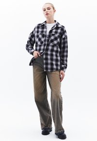 Svart och vit rutig flanellskjorta med två framskönt fickor, ihop med ljusbruna vida jeans och svart skor. Enkel bakgrund.