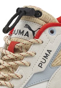 Beige und graue Sneaker mit strukturiertem Obermaterial, mehrfarbigen Schnürsenkeln und Puma-Logo. Enthält einen schwarzen Versteller für die Schnürsenkelanpassung.