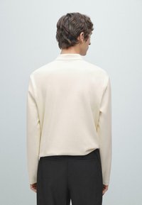 Mężczyzna z krótkimi brązowymi włosami, zwrócony tyłem, ubrany w kremową koszulę z długim rękawem i czarne spodnie, na jednolitym jasnoszarym tle.