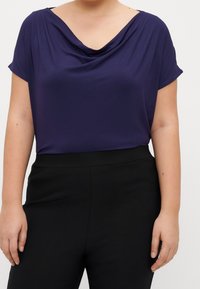 Marinblå kortärmad topp med en cowl-halsringning, mjukt tyg, draperad design, tillsammans med svarta högmidjade byxor.