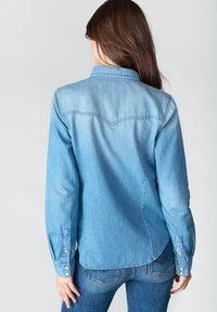 Chemise en denim bleu clair avec un col pointu, des manches longues et des fermetures à pression. Présente des détails de empiècement et un ourlet incurvé.