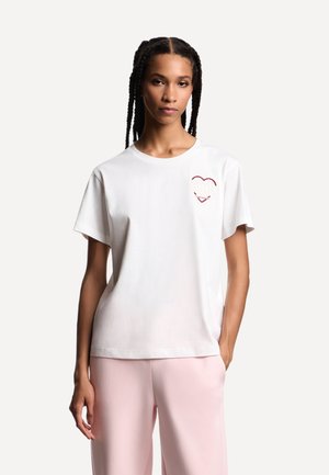 T-shirt bianca in cotone con maniche corte, con un disegno di cuore rosso e sorriso sul petto a sinistra. Tessuto morbido, vestibilità rilassata.