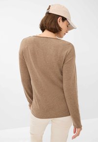 Brauner, langärmeliger Pullover aus weichem Material, mit lockerer Passform und gerundetem Saum. Kombiniert mit hellen, gerippten Hosen und einer beigen Mütze.