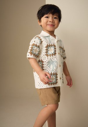 Băiat tânăr care zâmbește, purtând un tricou cu mâneci scurte croșetat cu modele albastre, maronii și albe și pantaloni scurți bej, stând în fața unui fundal simplu.