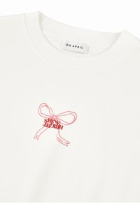 Felpa bianca con collo a costine rotondo, caratterizzata da una grafica con fiocco rosa e la scritta "SHINE BRIGHT" sotto. L'etichetta recita "OH APRIL."