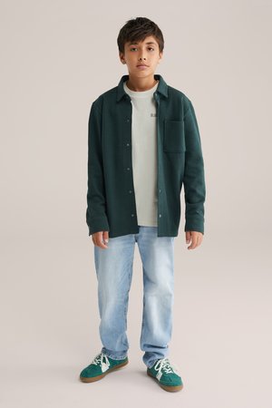 Grön skjorta med knappar och ficka, lagd över en ljusgrön t-shirt; matchad med ljusblå jeans och gröna sneakers.