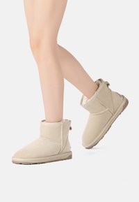 Beige suede ankle boots with a soft, fur-lined interior and a flat rubber sole. Feature stitched accents and a rear pull tab for easy wear.