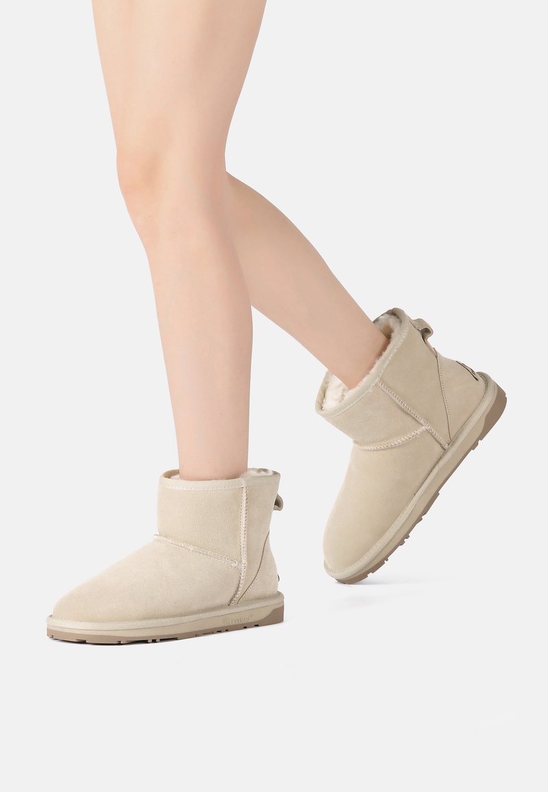 Beige suede ankle boots with a soft, fur-lined interior and a flat rubber sole. Feature stitched accents and a rear pull tab for easy wear.
