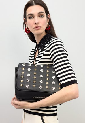 Bolso de mano de cuero negro con tachuelas metálicas y adornos transparentes, que presenta una forma estructurada y un logo en la parte inferior. Parte superior a rayas.