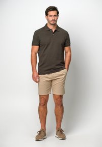 Polo shirt marron foncé à manches courtes, associé à un short beige. Le modèle porte des baskets marron clair et se tient devant un fond neutre.