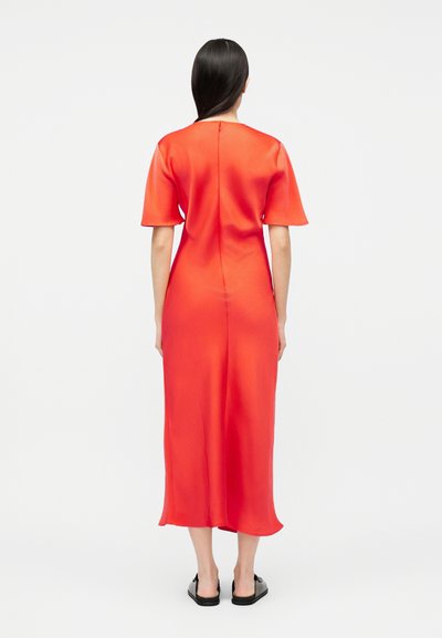 Γυναίκα με μακριά σκούρα μαλλιά που φοράει φωτεινό πορτοκαλί midi φόρεμα και μαύρα επίπεδα παπούτσια, στέκεται γυρισμένη με την πλάτη σε απλό λευκό φόντο.