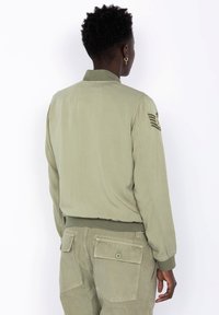 Khaki bomber jacket s rebrovaným golierom a manžetami. Má nášivku na pleci a hladkú, ľahkú textúru.